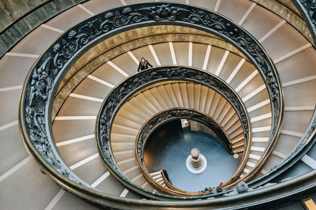 A figure paused in their ascent of an ornate spiral staircase, representing the stages of business growth and the owner who must grow to match each one.