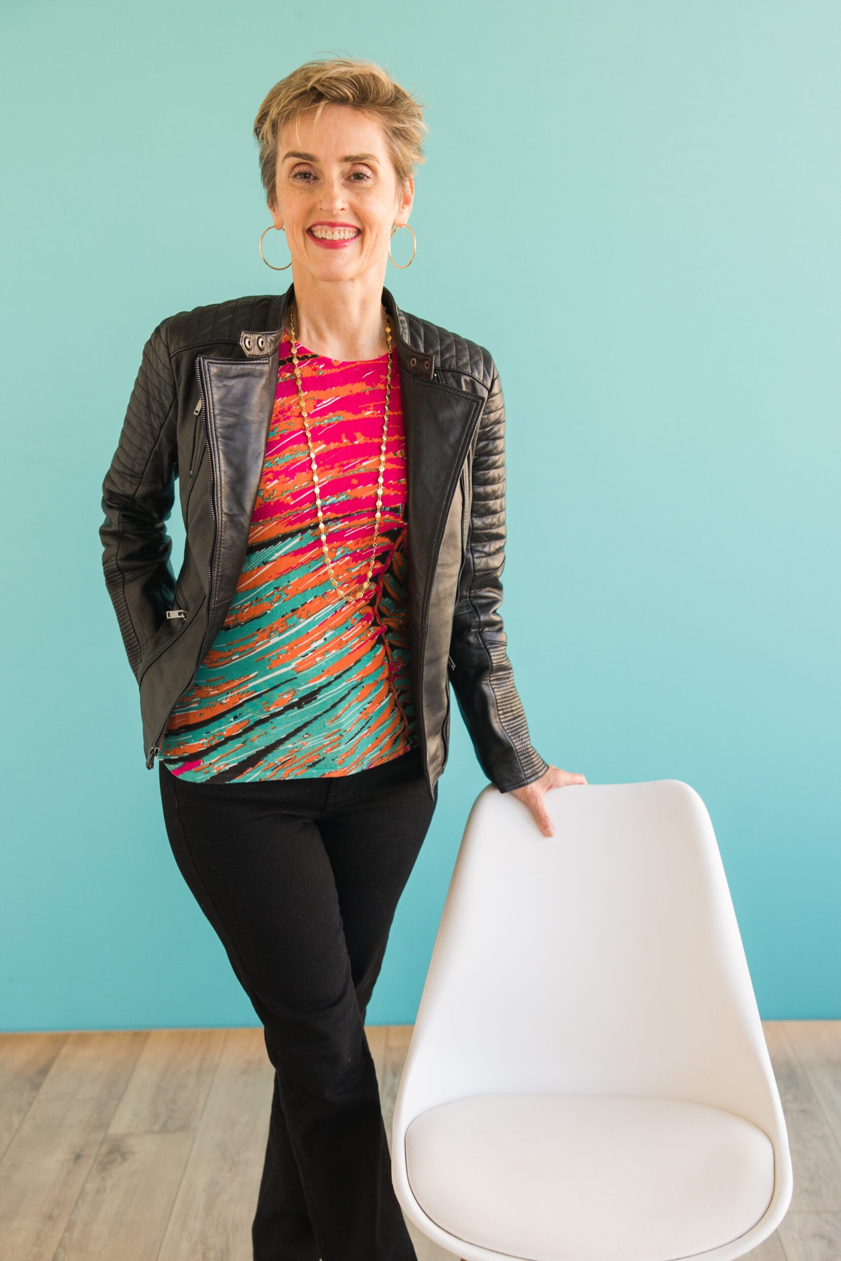 Karen Finn, PhD, business strategist, smiling and leaning on a white chair against a teal wall
